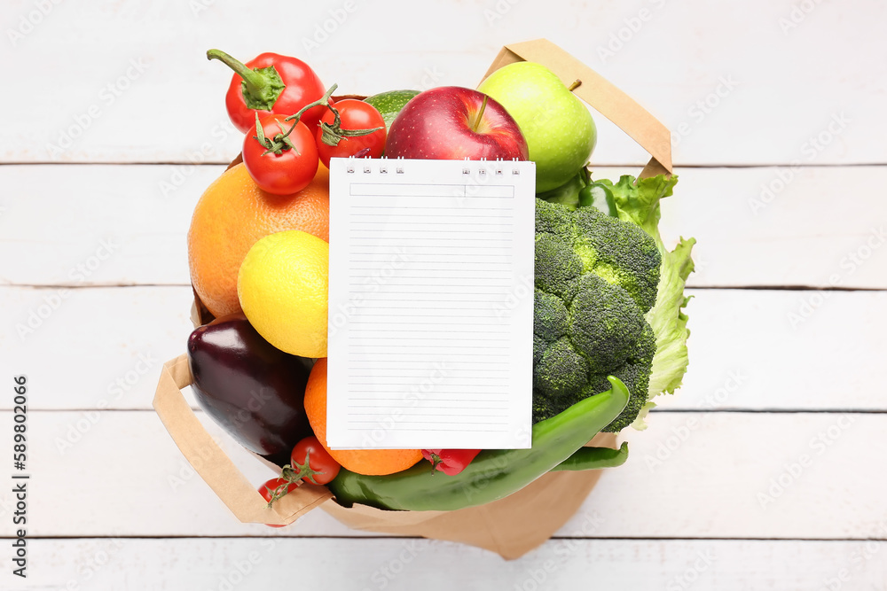 Paper bag with vegetables, fruits and blank notebook on white wooden background