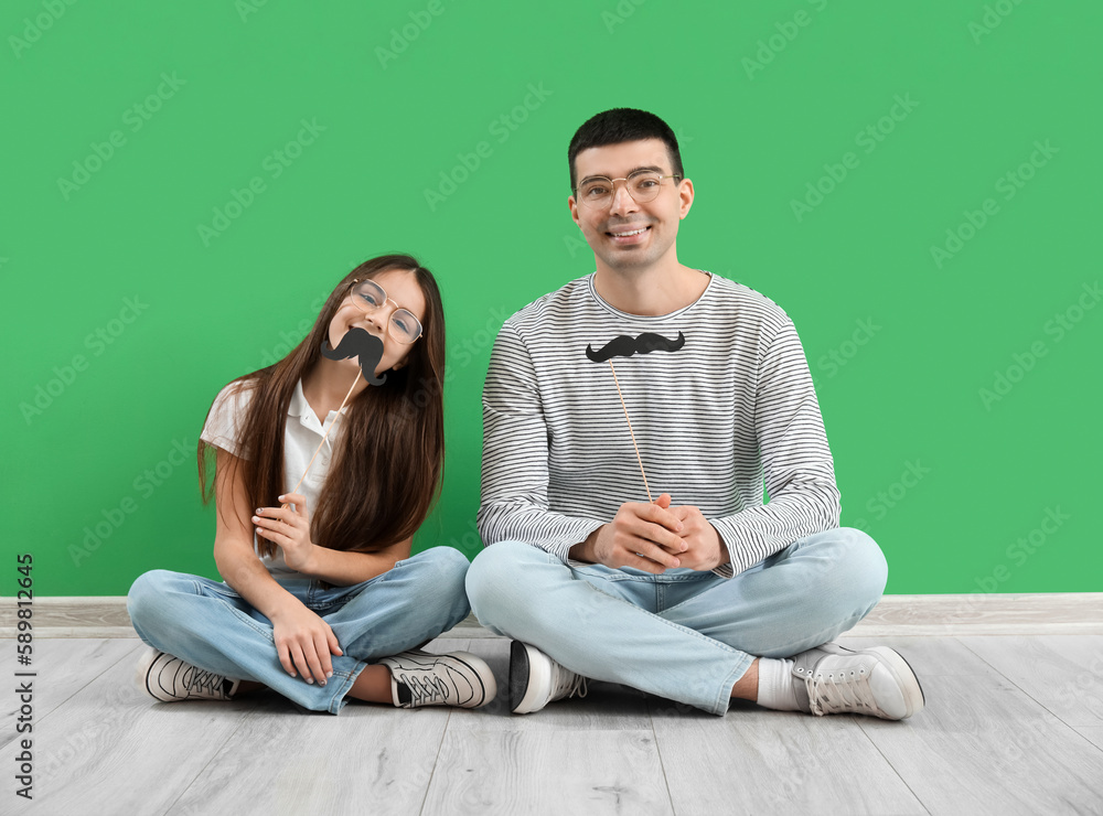 Portrait of father and his little daughter with paper mustache sitting near green wall