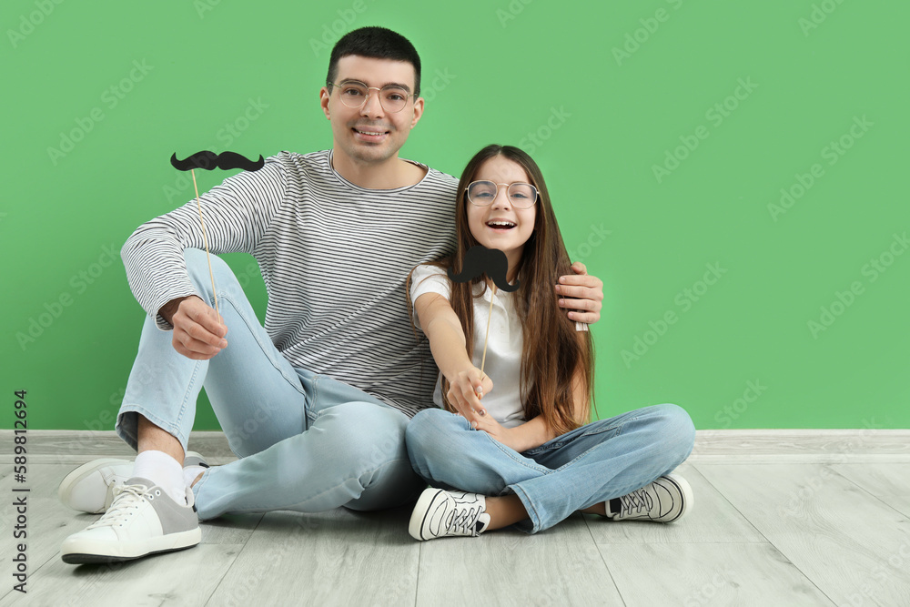 Portrait of father and his little daughter with paper mustache sitting near green wall