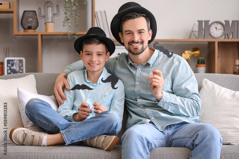 Portrait of father and his little son with paper mustache sitting on sofa