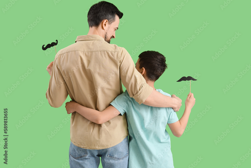 Father and his little son with paper mustache hugging on green background, back view