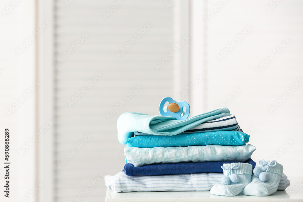 Stack of baby clothes and pacifier on table near folding screen