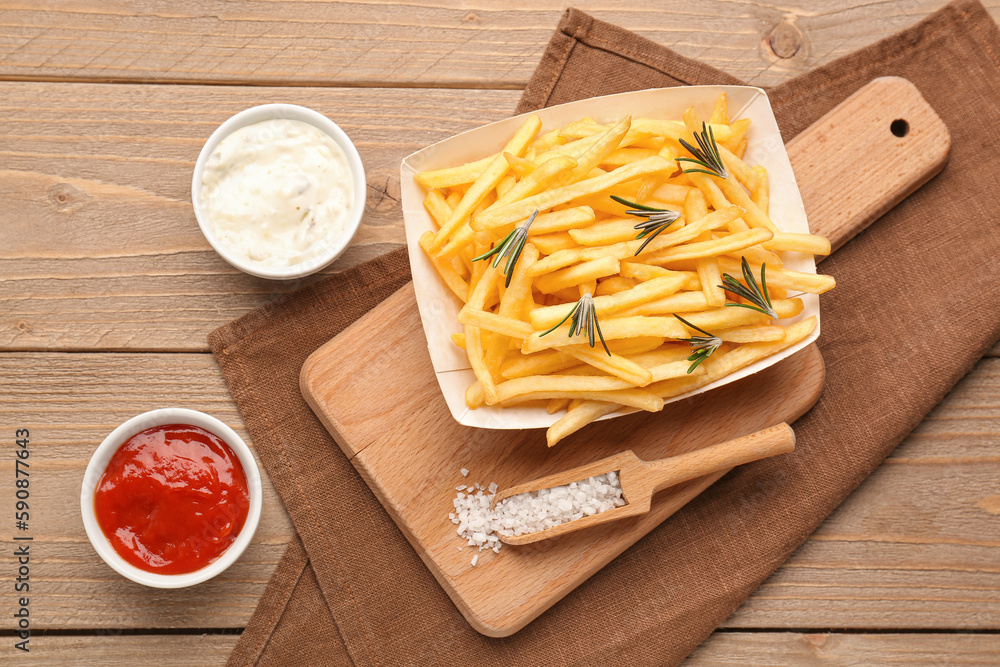 Paper box with tasty french fries, salt and sauces on wooden background