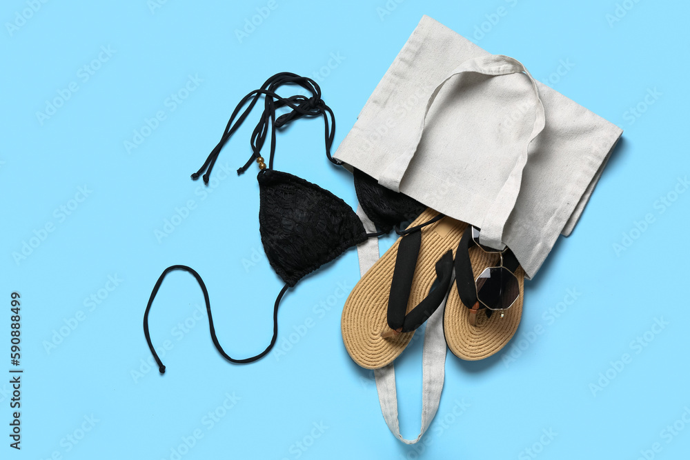 Bag with flip-flops, sunglasses and swimsuit on blue background