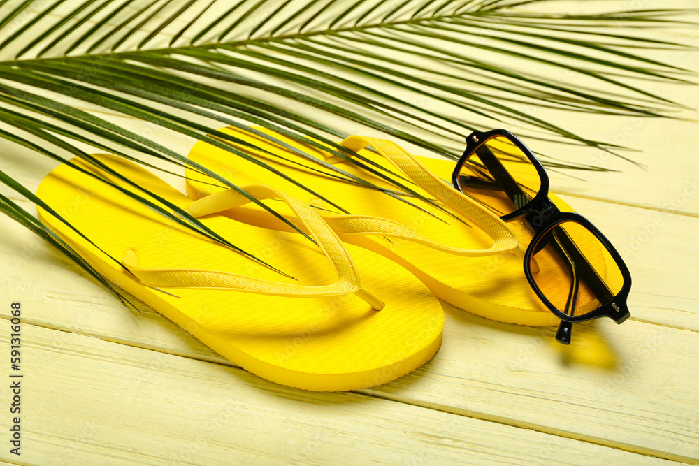 Flip-flops with sunglasses and palm leaf on yellow wooden background, closeup