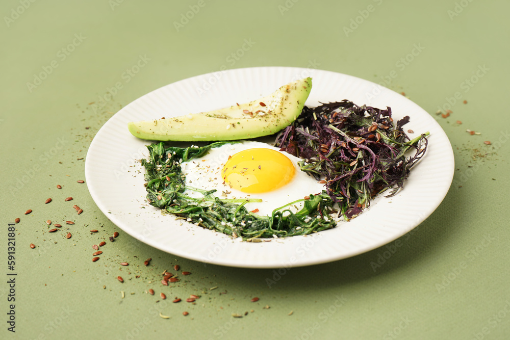 Plate with tasty fried egg, avocado and greens on green background