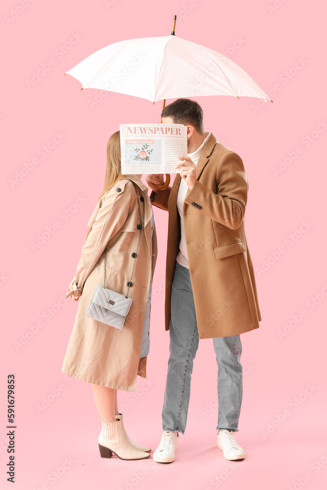 Young couple with umbrella kissing behind newspaper on pink background