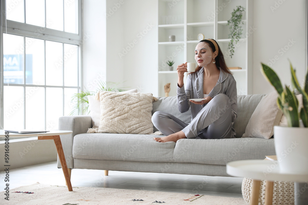 Young woman with sheet mask and cup of coffee sitting on sofa at home