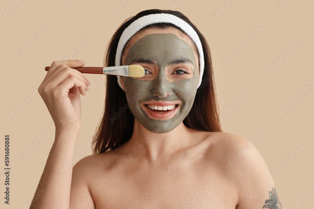 Young woman with applied clay mask and brush on beige background, closeup