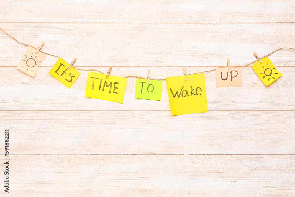 Sticky notes with text ITS TIME TO WAKE UP hanging on rope against light wooden background