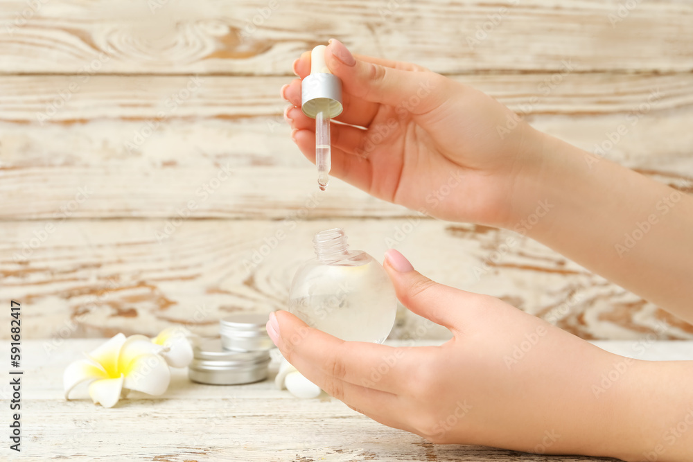 Female hands with bottle of cuticle oil, cosmetics and flowers on light wooden background, closeup