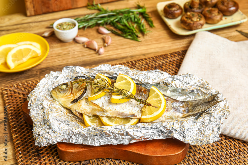 Aluminium foil with tasty baked fish on wooden table in kitchen