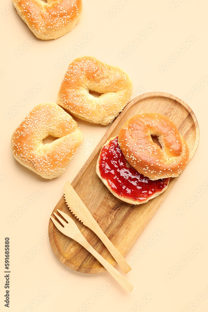 Wooden board of tasty bagels with sesame seeds and strawberry jam on beige background