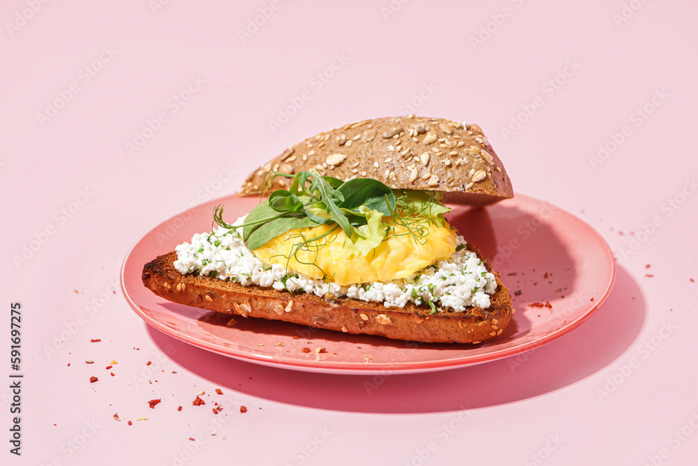 Plate with tasty scrambled eggs sandwich and greens on pink background