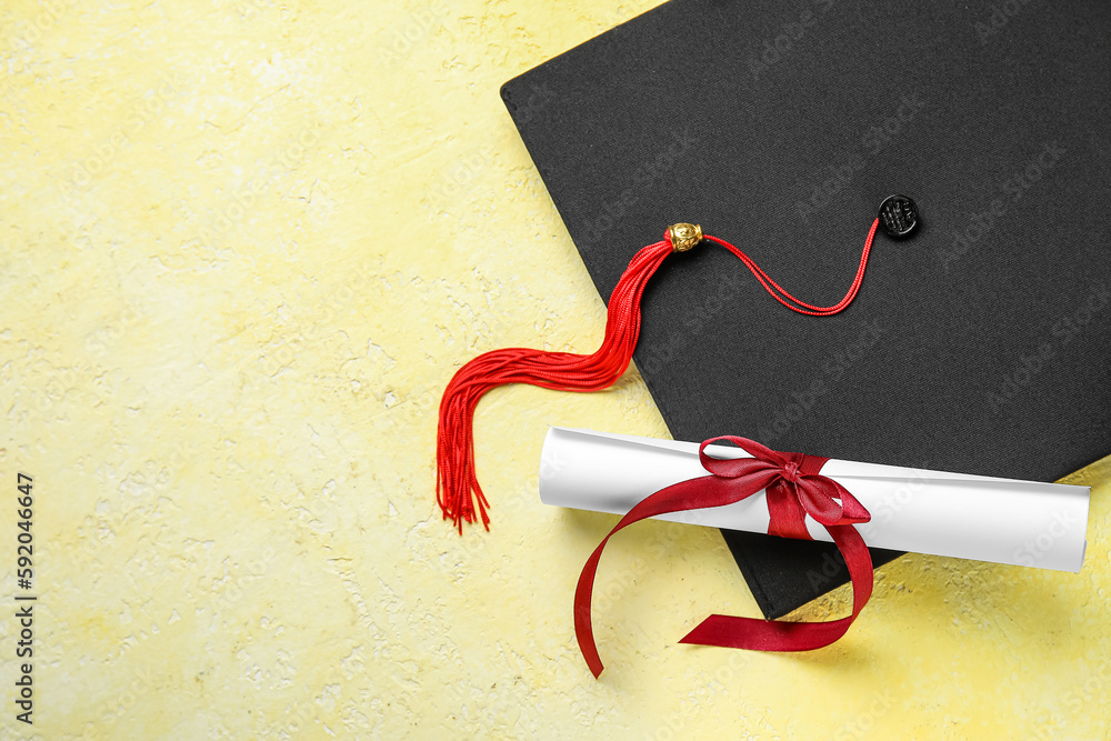 Diploma with red ribbon and graduation hat on yellow table