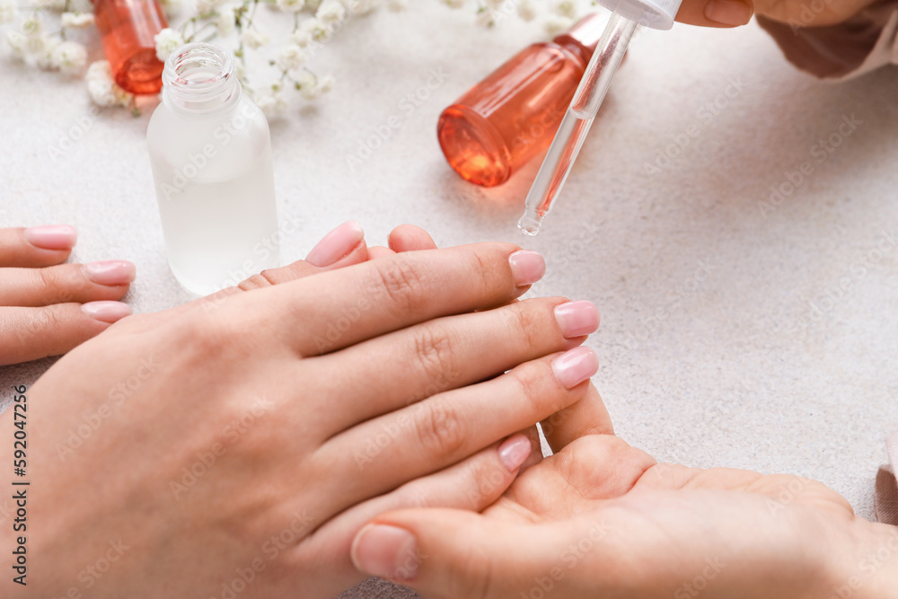 Manicure master applying cuticle oil onto female fingernails on light background, closeup