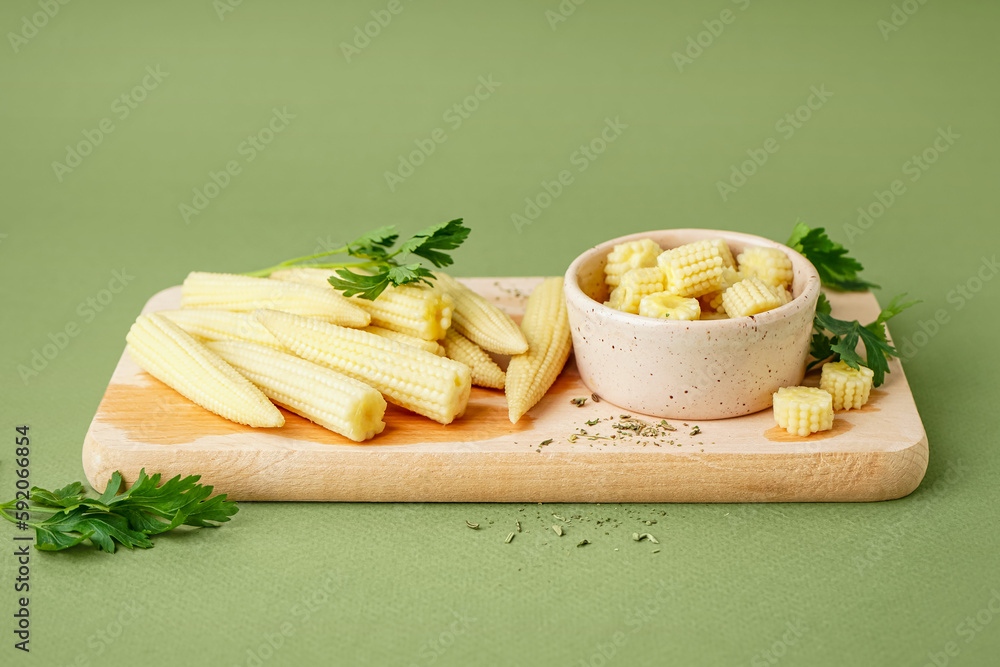 Wooden board with cut and whole canned baby corn cobs on olive background