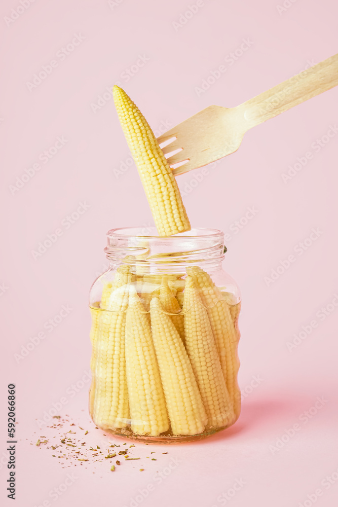 Jar and fork with canned baby corn cobs on pink background