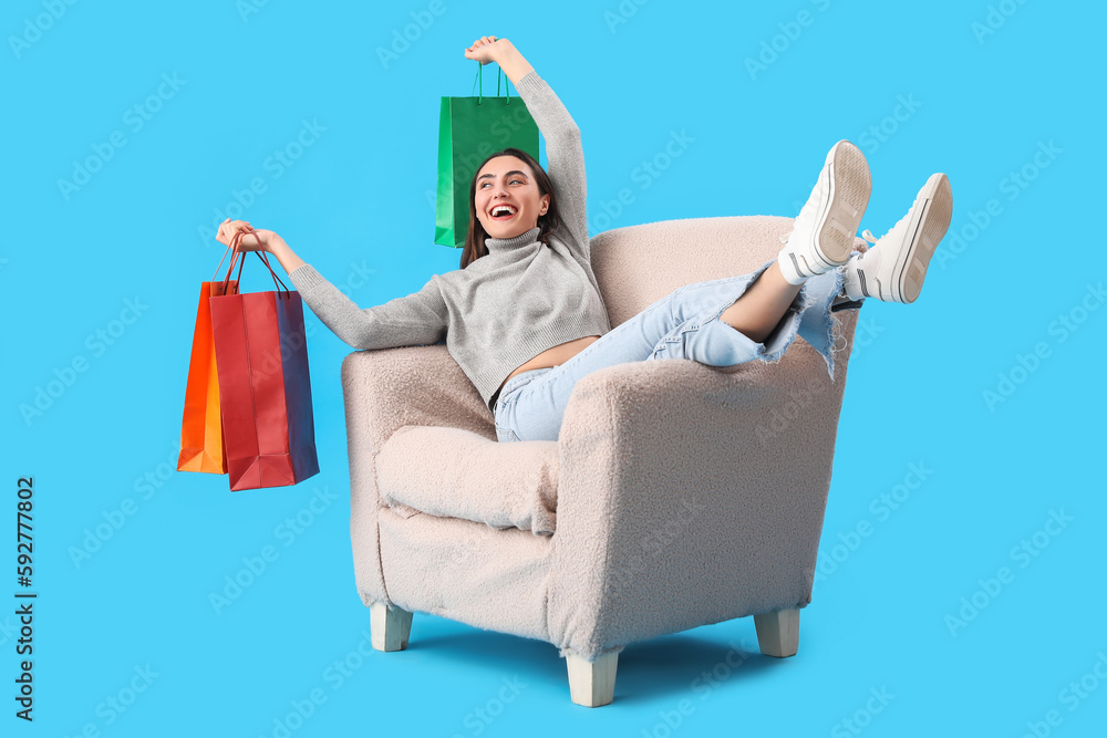 Young woman with shopping bags sitting in soft armchair on blue background