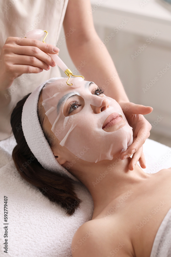 Cosmetologist massaging womans face with sheet mask in salon, closeup