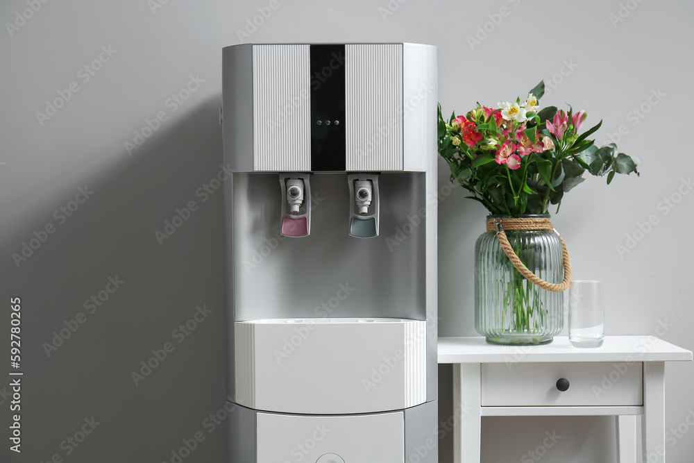 Modern water cooler and vase with beautiful flowers on end table on grey background