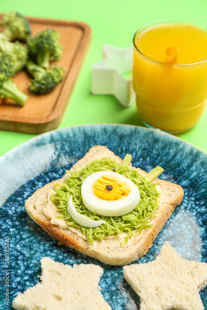 Plate with funny breakfast in shape of alien, vegetables and glass of juice on green background