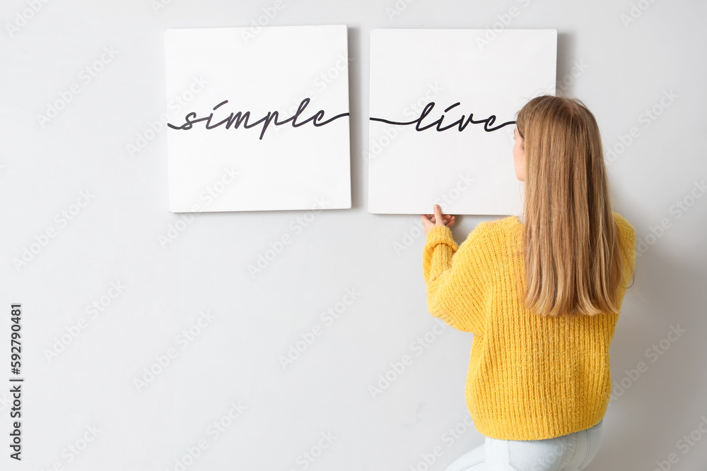 Young woman hanging poster on light wall at home