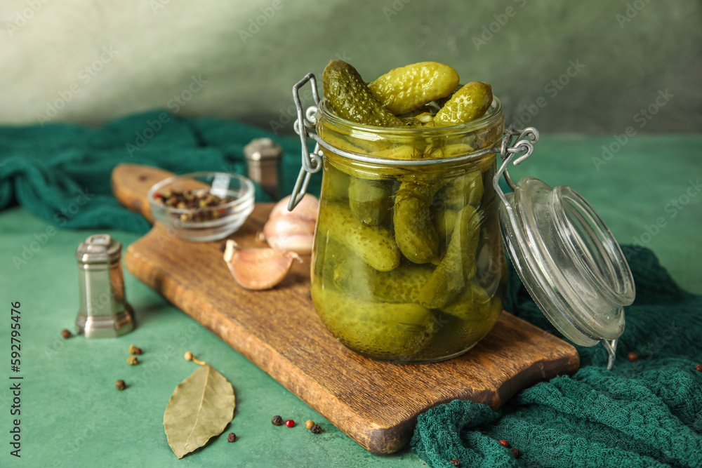 Jar with tasty fermented cucumbers, wooden board and ingredients on green table