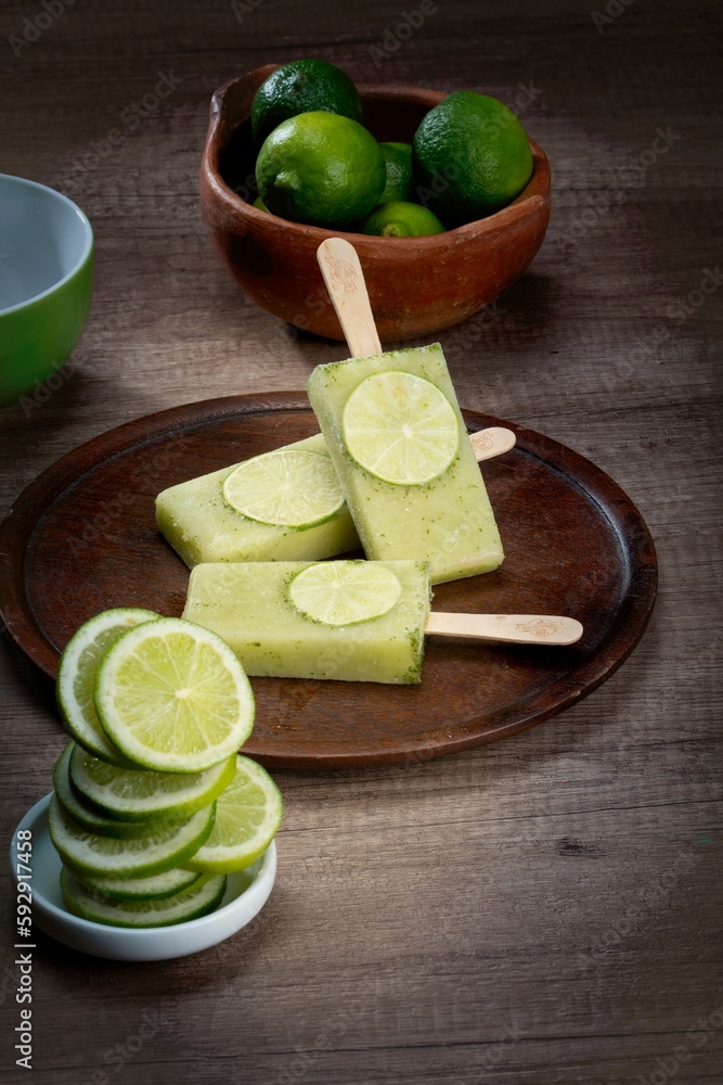 Vertical shot of lime flavor popsicles on a plate surrounded by full and sliced limes