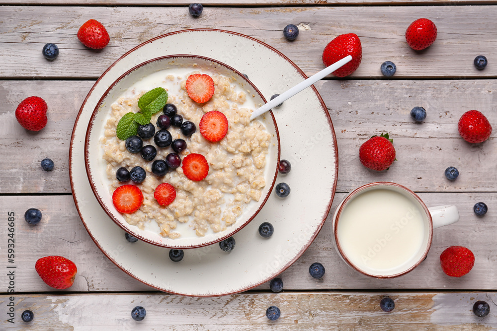 Bowl with tasty oatmeal, blueberries, strawberries and cup of milk on wooden background