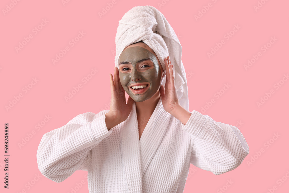 Young woman in bathrobe with applied clay mask on pink background