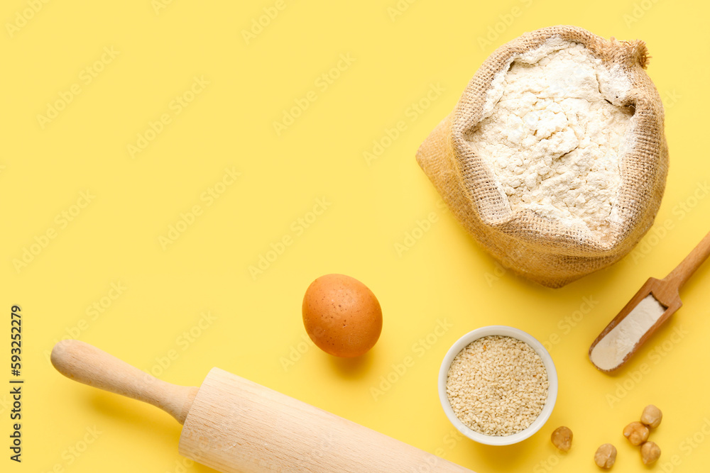 Composition with sack bag of wheat flour, sesame seeds, egg and rolling pin on yellow background