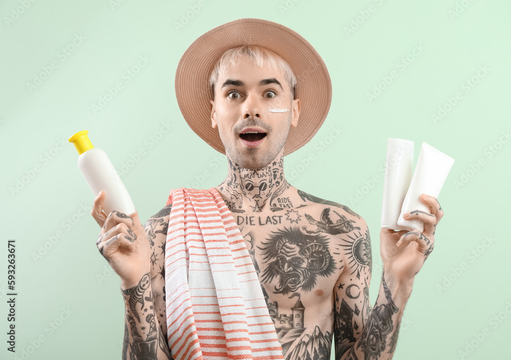 Surprised tattooed man with sunscreen creams on light green background