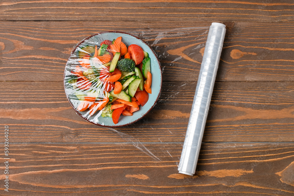 Bowl with salad wrapped with stretch wrap on wooden background