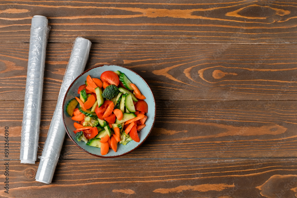 Bowl with salad and rolls of stretch wrap on wooden background