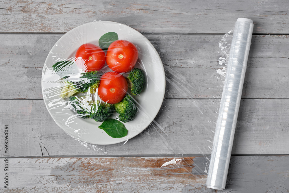 Plate with vegetables wrapped with stretch wrap on grey wooden background