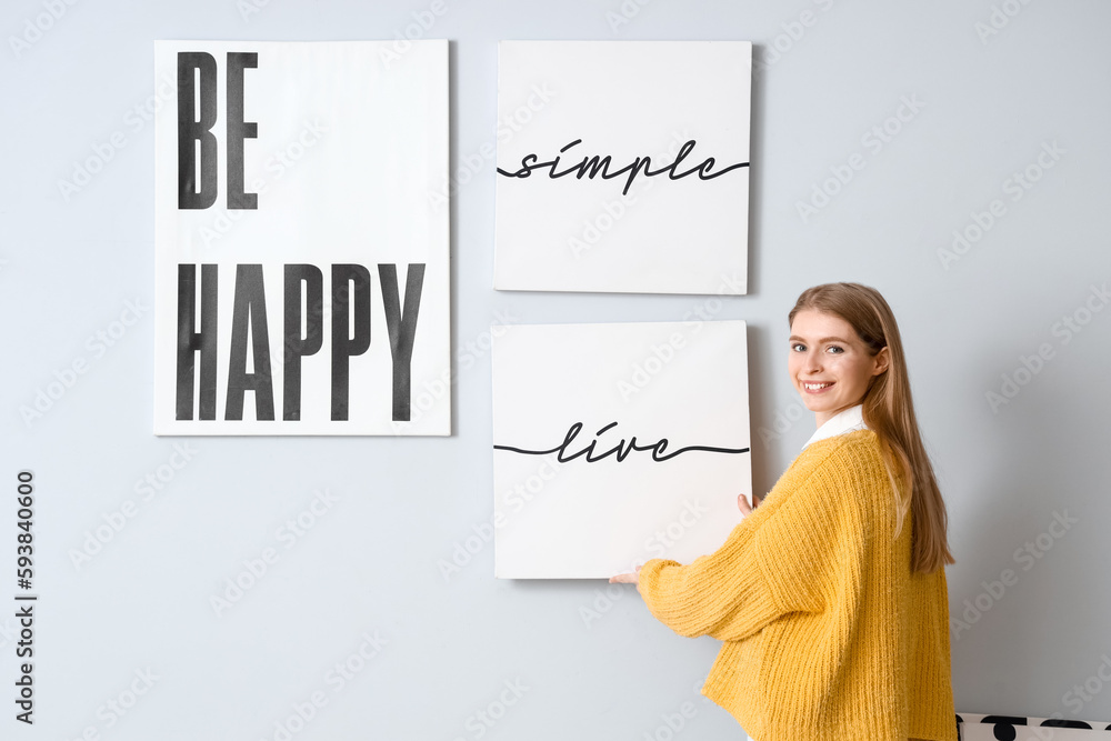 Young woman hanging poster on light wall at home