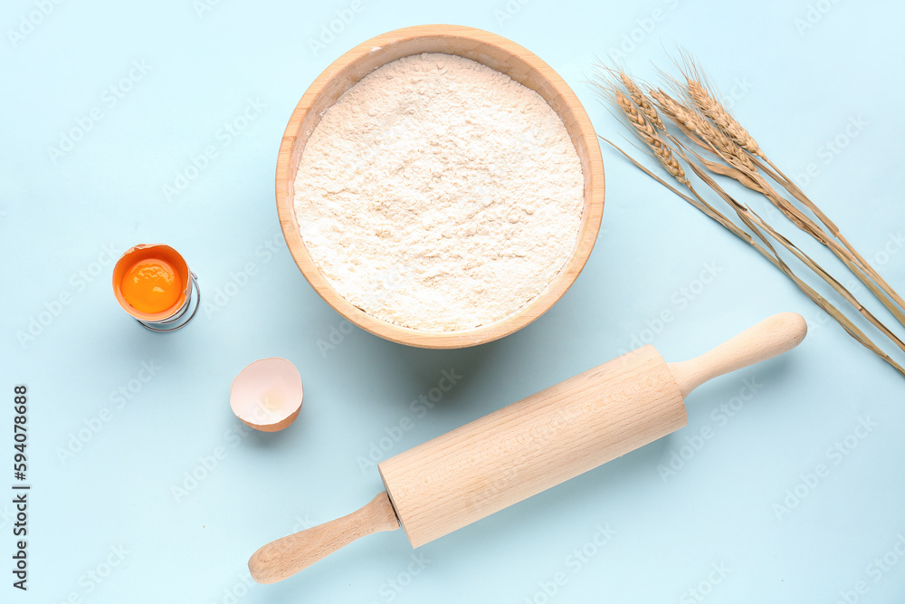 Bowl with flour, wheat ears, cracked egg and rolling pin on blue background