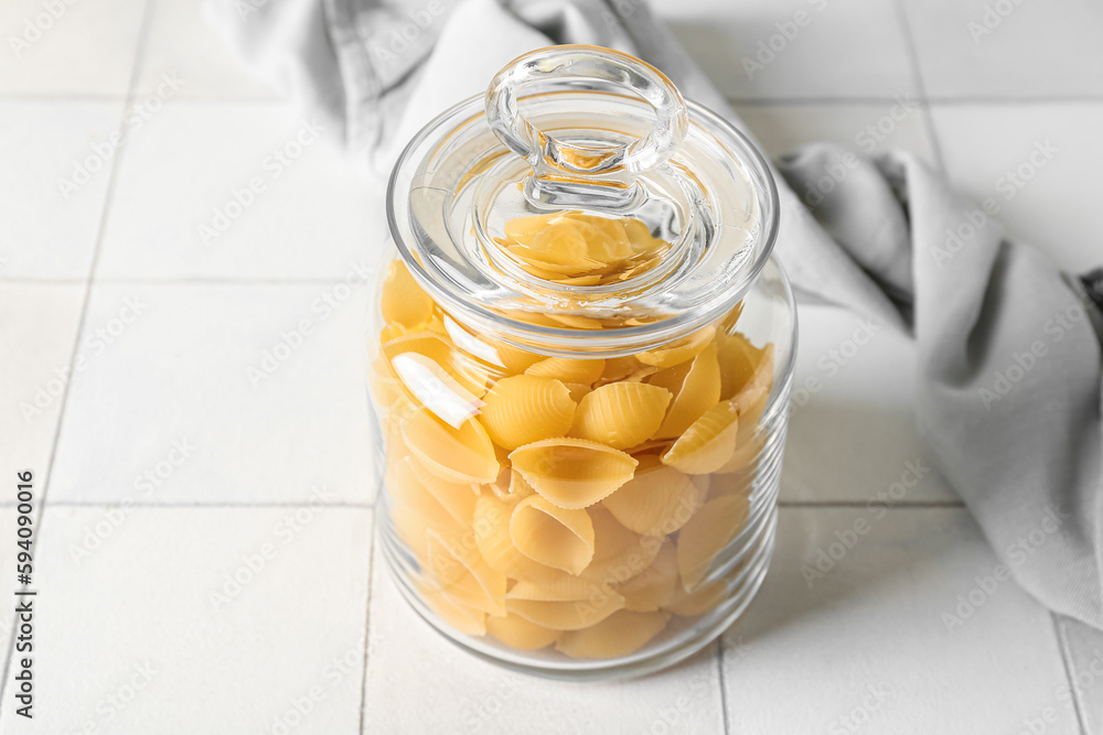 Jar with raw conchiglie pasta on white tile background