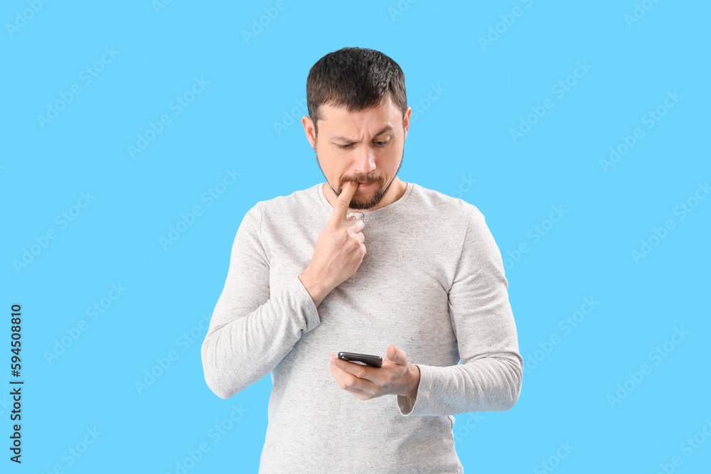 Handsome man with mobile phone biting nails on blue background