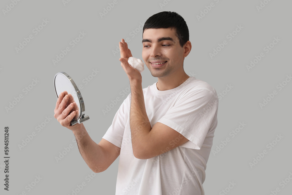 Young man applying shaving foam onto face against light background