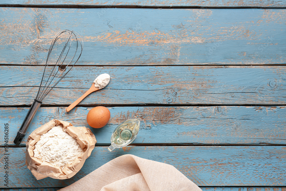 Bag of wheat flour, egg, oil, whisk and spoon on blue wooden table