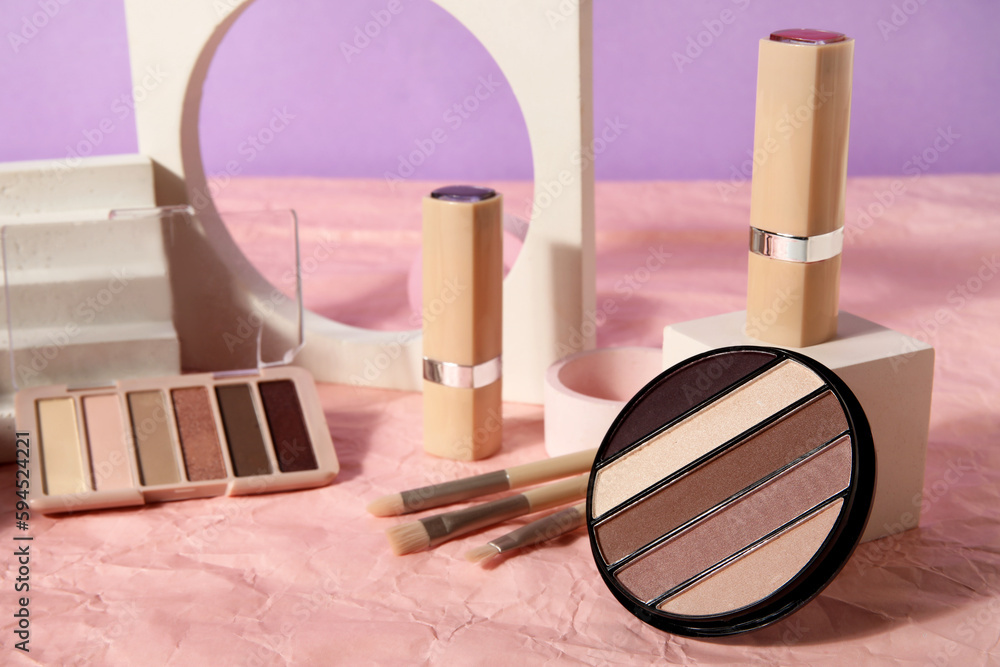 Decorative cosmetics with brushes and podiums on crumpled paper against lilac background, closeup