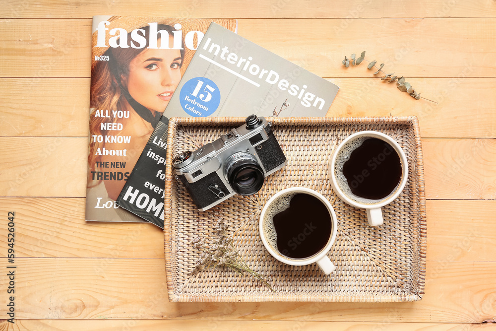 Tray with cups of coffee, photo camera and magazines on light wooden background