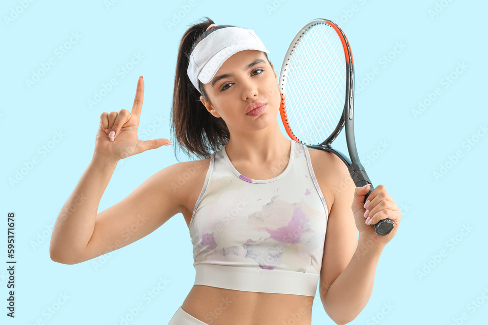 Sporty young woman with racket showing loser gesture on blue background