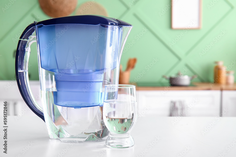 Modern filter jug and glass of water on table in kitchen