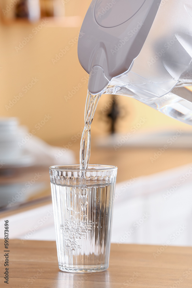 Pouring of water from modern filter jug into glass on kitchen counter