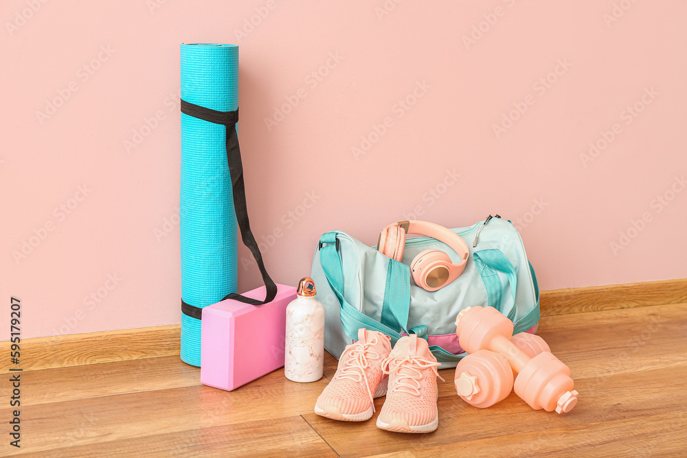 Sports bag with headphones, bottle, mat, sneakers and dumbbells near pink wall