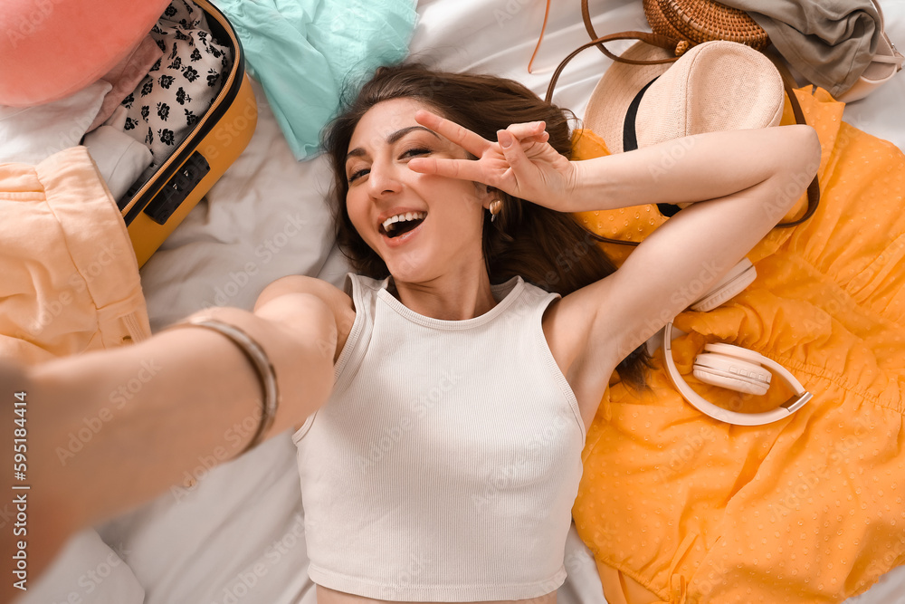 Young woman with beach accessories and suitcase taking selfie on bed