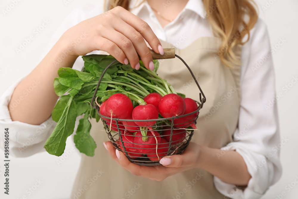 Woman holding basket of ripe radish with green leaves on light background, closeup